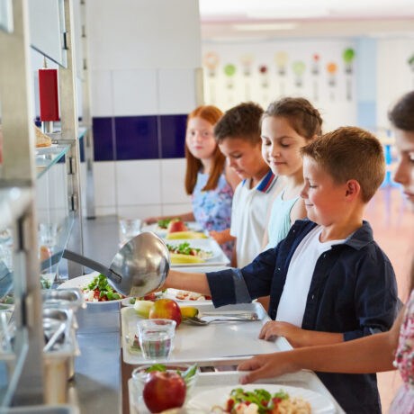 Una fila de niños en el comedor de un colegio sostienen bandejas mientras se sirven la comida en un mostrador de autoservicio. En primer plano, un niño con camisa azul oscuro utiliza un cucharón para servirse ensalada en su plato. El mostrador tiene estantes con manzanas rojas y vasos de agua, y al fondo se aprecian dibujos coloridos de cubiertos en las paredes del comedor.