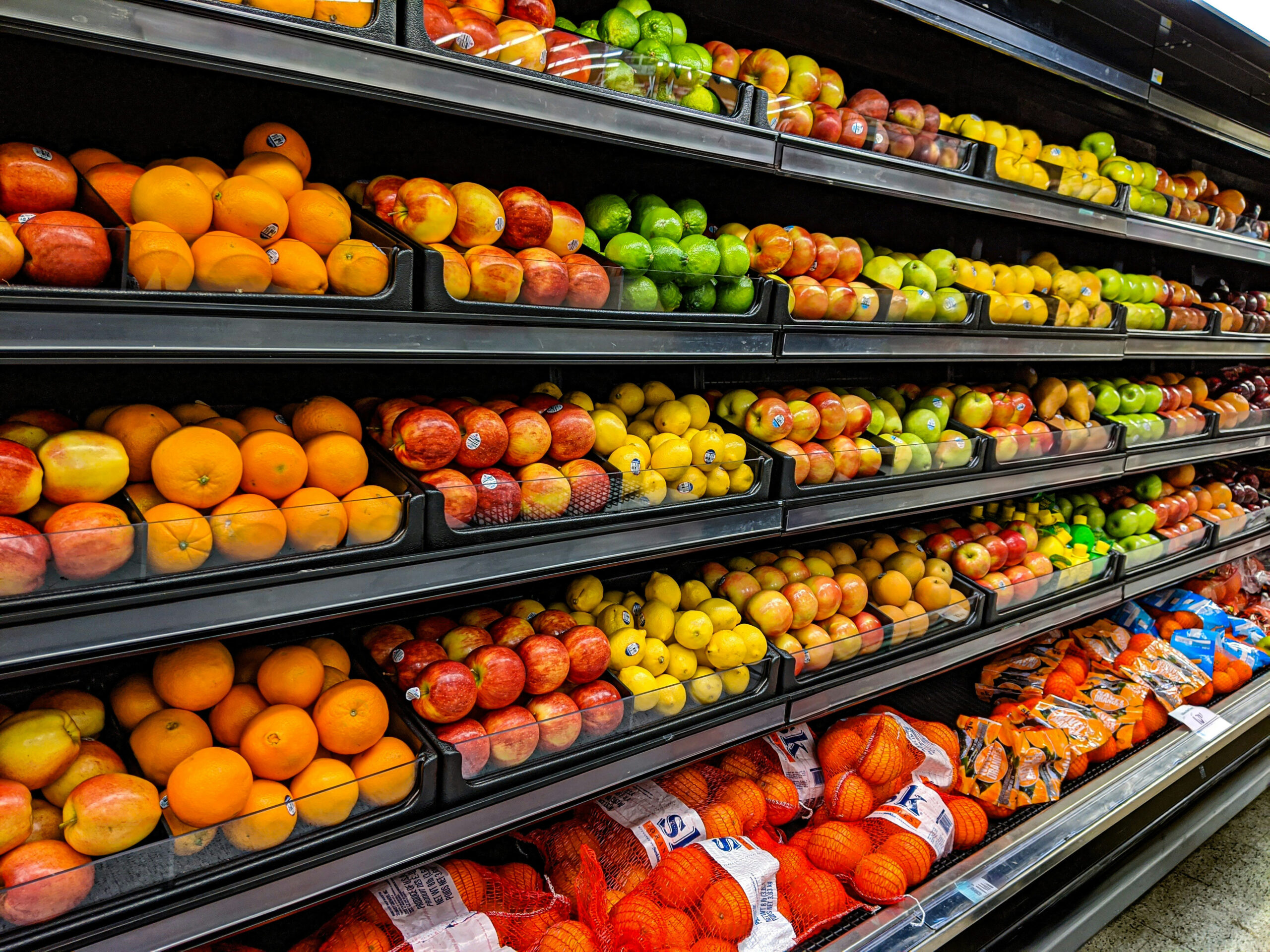 Sección de frutas de un supermercado con naranjas, manzanas, limones y otras frutas frescas organizadas en estanterías.