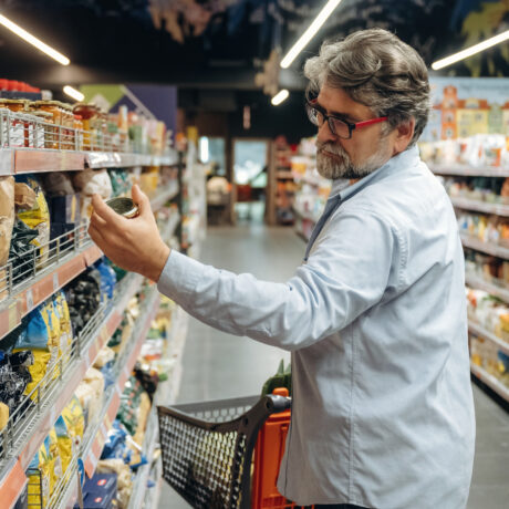 Persona realizando la compra en un supermercado, observando y leyendo la etiqueta de un producto envasado en uno de los pasillos. A ambos lados se ven estanterías repletas de alimentos y productos de consumo, mientras un carrito de la compra acompaña a la persona durante la selección de productos.