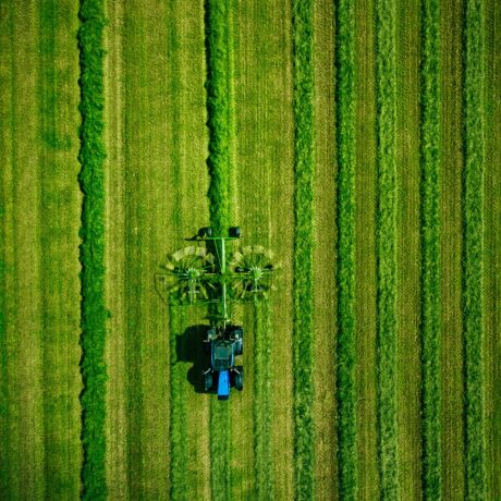 Vista aérea de un tractor con una máquina agrícola trabajando en un extenso campo verde, dejando franjas de vegetación diferenciadas a su paso. El terreno presenta largas hileras de cultivo en distintos tonos de verde.